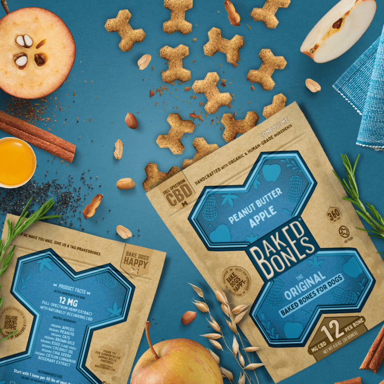 Busy image including golden brown baked dog bones pouring out of two Kraft and blue bags of BakedBones, cut apples, rosemary, shelled peanuts, cinnamon, and chia seeds, all against a blue background